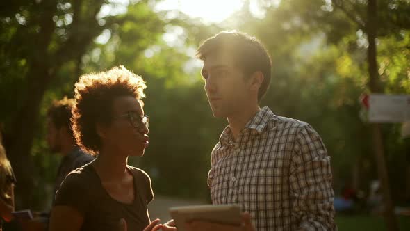 Darkskinned Female Standing Talking with Caucasian Young Male with Tablet in Park in Slowmotion alt