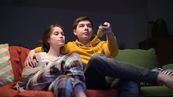 A young guy and his girlfriend sit on the sofa and turn on the TV in the evening alt