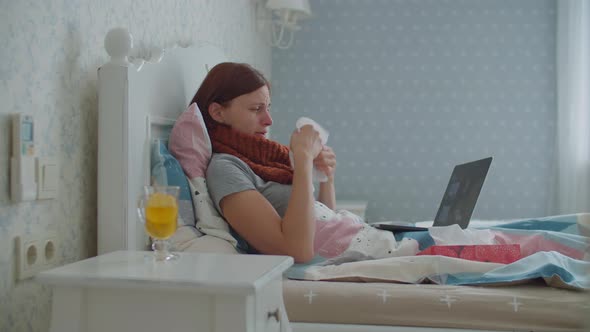 Sick Young Woman Laying in Bed and Talking Online By Laptop. Woman with Warm Scarf on Neck Coughs