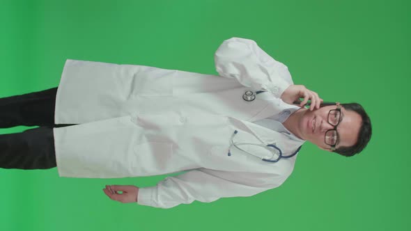 Asian Doctor With Stethoscope Talking On Mobile Phone While Walking On Green Screen alt
