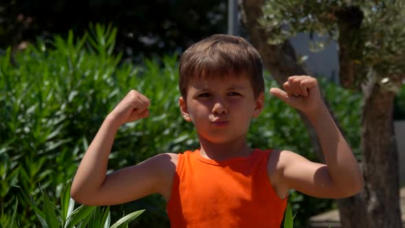 Little Serious Tanned Boy in the Orange Top is Showing His Biceps Muscle alt