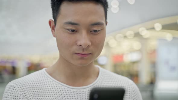 Close Up of Asian Male Scrolling Phone on Big Mall Background