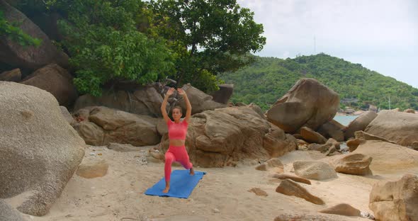Woman Exercising on Blue Mat She Makes Warrior One Exercise on the Small Beach Between the Rocks alt