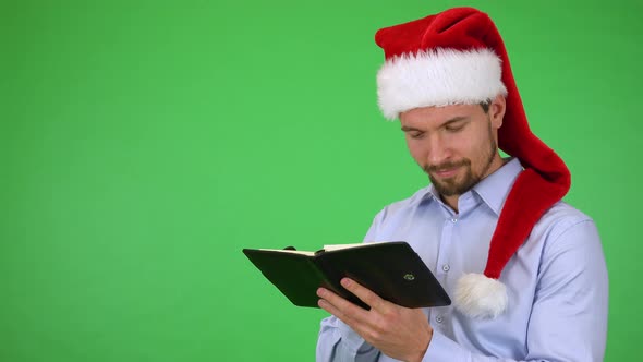 A Man in a Christmas Hat Writes a Wish List Into a Notebook with a Smile - Green Screen Studio alt