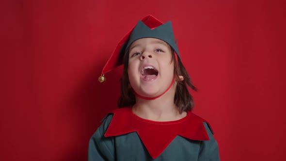 Baby Boy in a Green Suit and a Dwarf Hat is Indulging on Red Background for Christmas alt