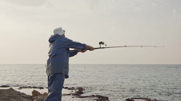 Old fisherman standing on sea side rocks and fishing against the sunset alt