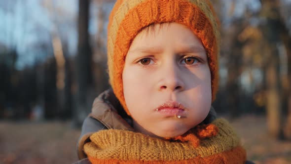 Boy in a Knitted Hat and a Snood Chewing Food with His Mouth Closed in an Autumn Park alt