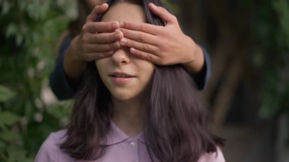 Closeup Portrait of Cheerful Teenage Girl in Park with Friend Covering Eyes with Hands alt