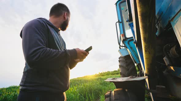 Man in Field with Phone in His Hands Stands By His Tractor and Knocks on Wheel Checking Condition of alt