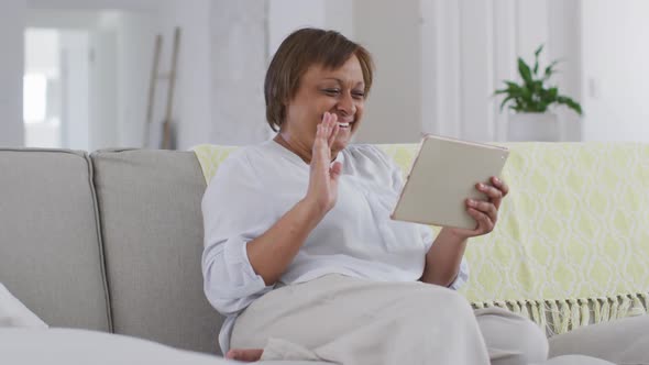Happy african american senior woman sitting on couch making video call using tablet, waving alt