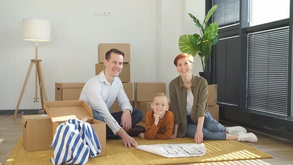 Happy Positive Family with Child Boy Sitting on Floor Among Cardboard Boxes in New House Using