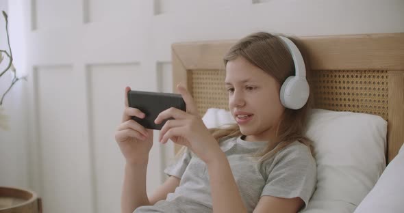 School Girl Is Playing Video Game in Smartphone By Internet, Using Headphones and Lying on Bed at alt