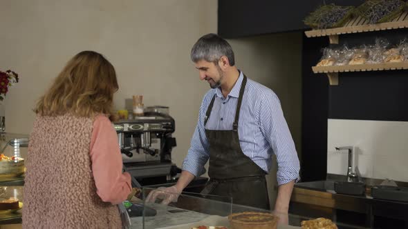 Handsome Male Cashier in Apron and Gloves Accepts Terminal Payment From Female Customer alt