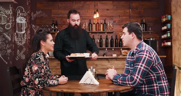 Bearded Waiter Bringing Cofffee and Croissants to a Gorgeous Inlove Couple alt