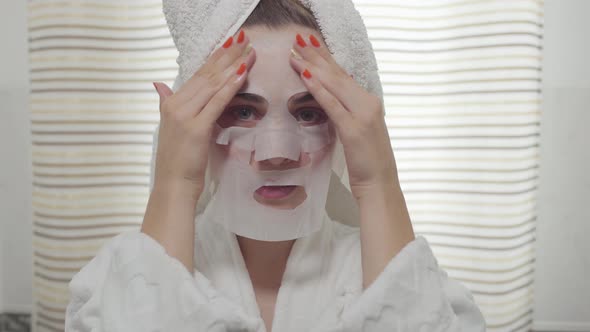 Cheerful Young Woman looking in the Camera Applying the Sheet Mask on Her Face in the Bathroom alt