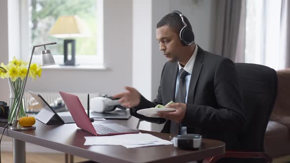 Young African American Man in Suit and Headphones Sitting in Home Office Talking in Video Chat and alt