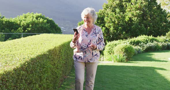 Caucasian senior woman smiling while talking on smartphone while walking in the garden alt