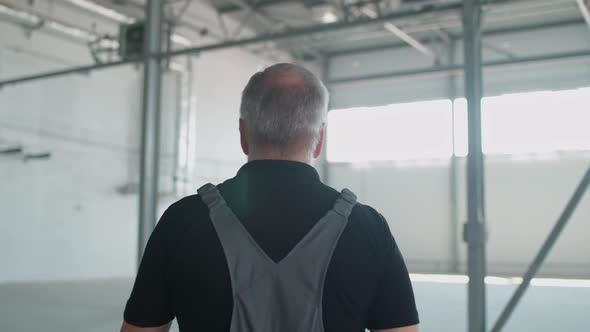 Back View of Grayhaired Man Worker Walks Through the Warehouse a Engineer Walks Through the alt