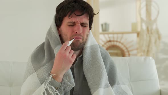 Man with a Cold is Sitting on the Couch with a Thermometer in His Mouth alt