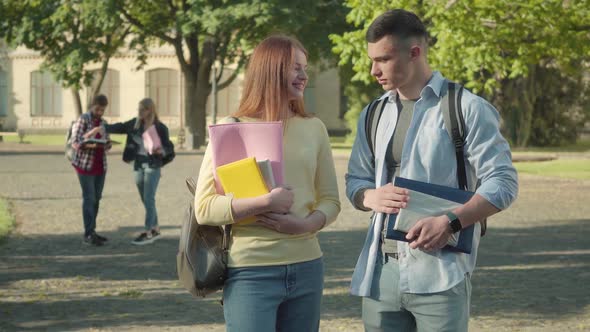 Portrait of Positive Caucasian Boy and Girl Talking on University Campus Yard with Blurred Students alt