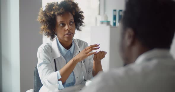 Africanamerican Woman Doctor Sitting at Table and Talking to Male Colleague in Office alt