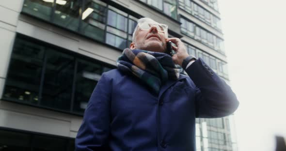 A Grayhaired Man Uses a Mobile Phone Standing in Business Center of the City alt