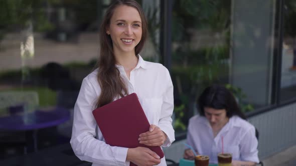 Portrait of Cute Confident Young Woman Posing Outdoors in Sidewalk Cafe with Blurred Friend Sitting alt