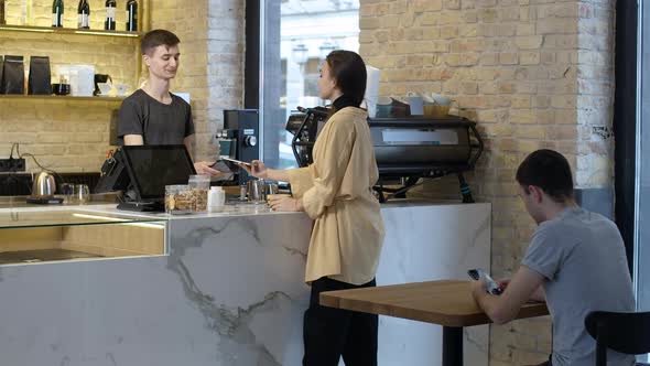 Wide Shot of Young Beautiful Woman Paying for Coffee Using Smartphone and Leaving with Cup alt