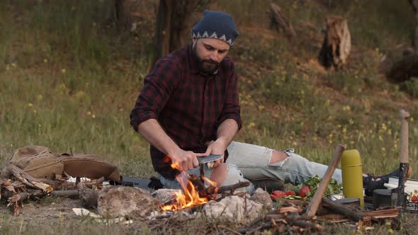 Tourist Sits Near Campfire and Sharpen a Stick with Knife