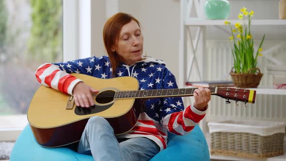 Talented Mature Woman Singing Playing Guitar Sitting on Bag Chair Indoors at Home alt