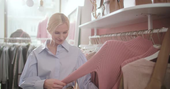 Young Attractive Blonde Woman Carefully Examines Clothes on a Rack or Hanger in a Shopping Center