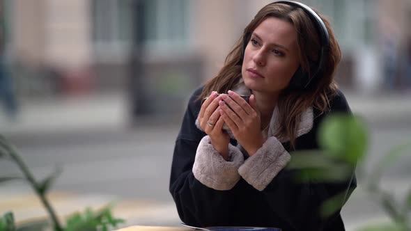 Portrait of a Sad Young Woman Listening To Music with Headphones in the Open Air. She Sits alt