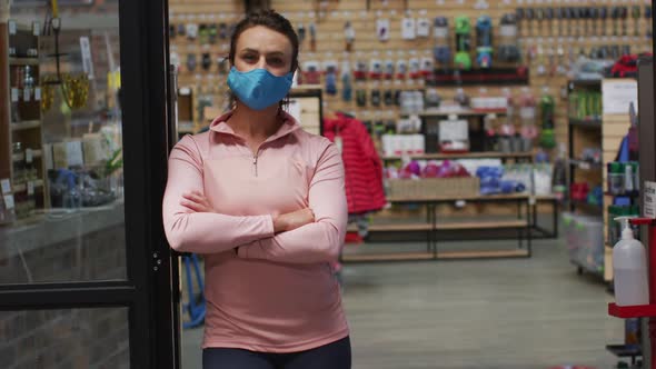 Caucasian female shopkeeper wearing face mask leaning in the doorway of sports shop alt