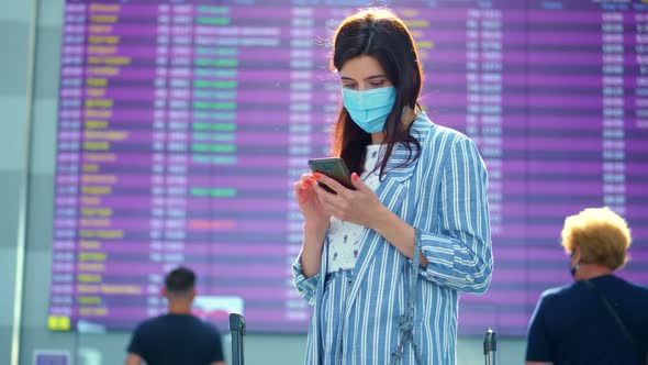 Female Air Passenger in Mask, Standing Against Departure Board at Airport. She Is Checking Flight alt