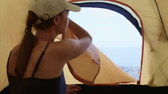 Woman unzips the tent inside and looks at sea stretching her arms in morning.