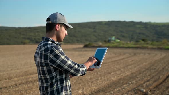 Farmer with Digital Tablet on a Background of Tractor Plowing in the Field alt