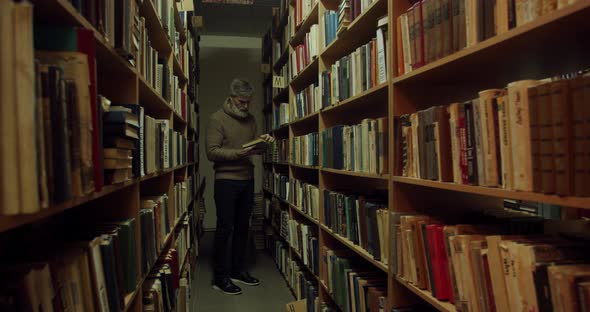 Old man is walking in an aisle of the library, looking at the books, 4k alt