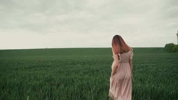 Young pregnant woman with flowing hair in a pink summer dress walks forward on a green field 
