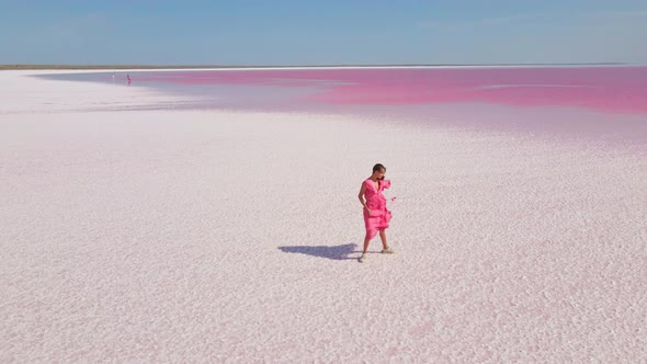 Cinematic Aerial Drone Following Attractive Young Woman in Pink Dress Walking on Colorful Pink Lake alt
