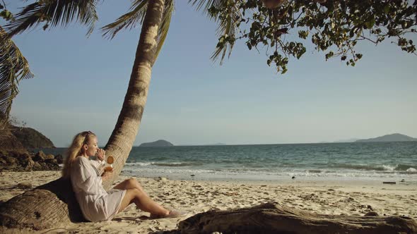 Woman Sitting in White Dress Drinks Pineapple Cocktail Pina Colada on Beach Near Palm alt