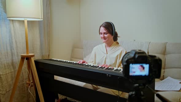 Woman musician with piano looking at video camera at home on sofa in living room alt