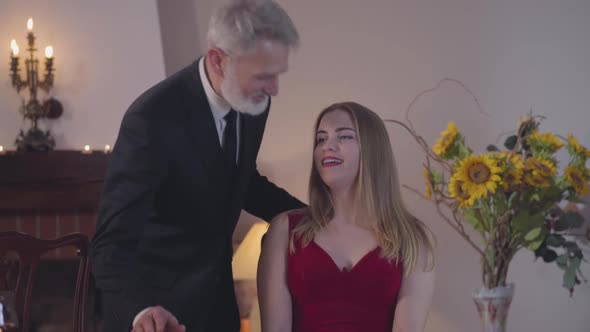 Elegant Middle-aged Caucasian Man Helping Beautiful Woman To Sit Down on the Chair. Graceful Pretty alt