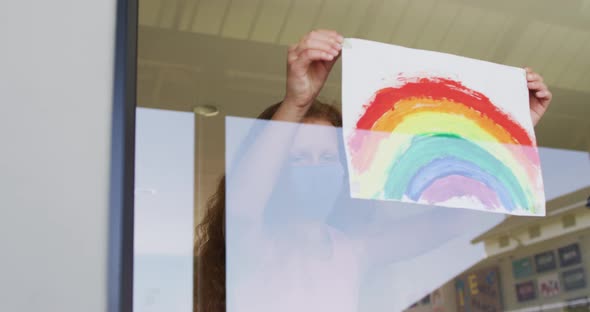 Girl wearing face mask sticking rainbow painting to the window at school alt