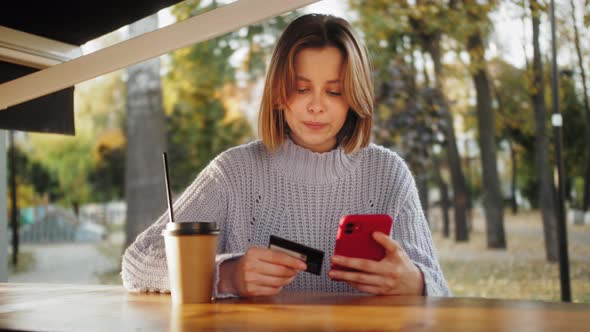 Smiling Young Customer Holding Credit Card and Smartphone Sitting in the Street Cafe alt