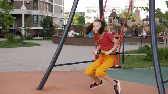 A Girl Rides on a Swing on the Background of a Playground in the Courtyard of a Residential Area alt