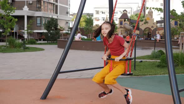 A Girl Rides on a Swing on the Background of a Playground in the Courtyard of a Residential Area