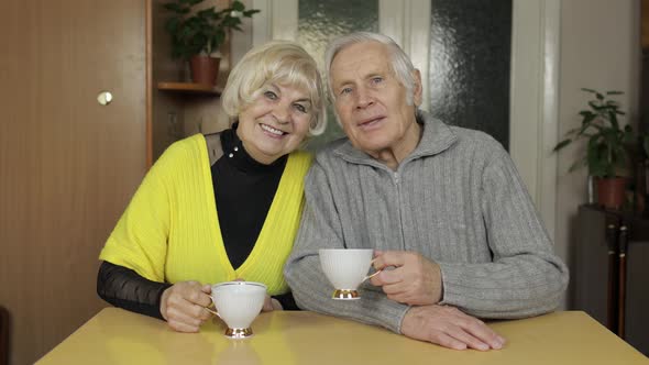 Happy Old Grandparents Couple Sit on Table at Home Enjoy Drinking Tea Together alt