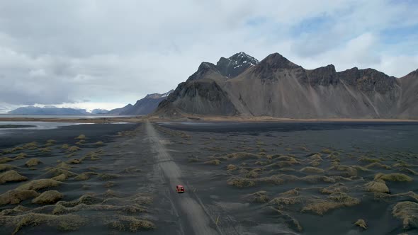 Drone video of Stokksnes Vestrahorn black sand beach in Iceland with Red car driving down dirt road alt