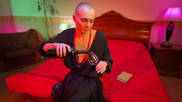 Young Man Pouring Wine in Glass Sitting on Bed in Red Lights alt