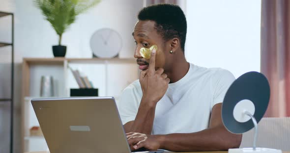 Black-Skinned Guy with Golden Eye-Patches Sitting at the Table at Home and Working on Computer alt